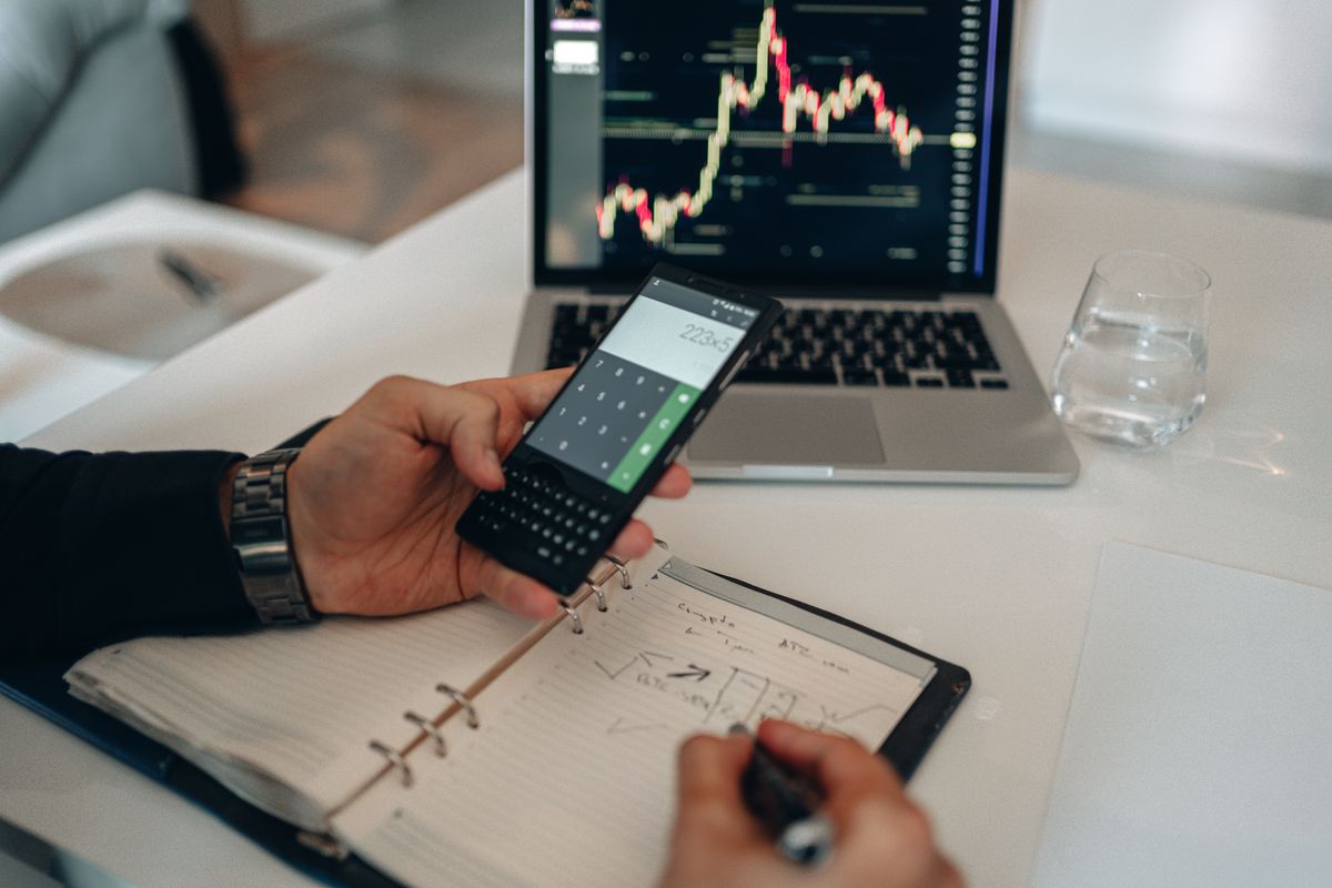 A hand working out a figure on a phone calculator beside a handwritten notebook and a laptop